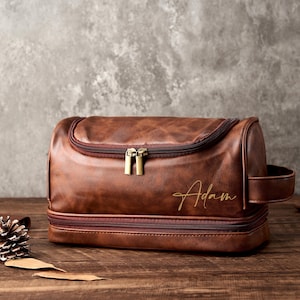 May include: A brown leather wash bag with a top and bottom zip. The bag has a side handle and the name "Adam" is written in gold script on the front. It is on a wooden surface.