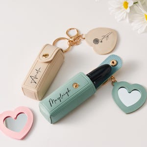 May include: Two rectangular lipstick cases with gold snap closures and gold keychains. One case is beige with the name Amelia, the other is sage green with the name Mayleigh. Each includes a heart-shaped charm, one with a mirror and one with a flower.