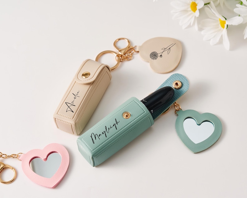 May include: Two rectangular lipstick cases with gold snap closures and gold keychains. One case is beige with the name Amelia, the other is sage green with the name Mayleigh. Each includes a heart-shaped charm, one with a mirror and one with a flower.