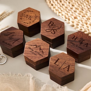 May include: A collection of six hexagonal wooden ring boxes with engraved initials and dates. The boxes are dark brown with lighter wood tops. Two silver rings are visible near the boxes. The boxes are personalized with initials and names.