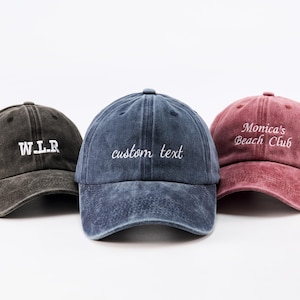 Può includere: Tre cappellini da baseball in diversi colori: grigio scuro, blu e rosso. Ogni cappellino ha un testo ricamato: "W.L.R", "custom text" e "Monica's Beach Club". I cappellini sono realizzati in un materiale di cotone lavato.