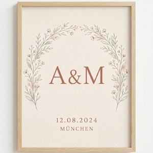 May include: A framed print featuring a light beige background with a floral arch made of thin brown stems and small pink flowers. The center displays the letters A&M in brown, with the date 12.08.2024 and the word MÜNCHEN printed below.
