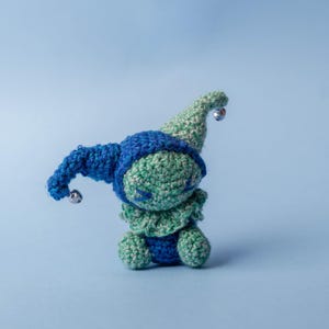 May include: A handmade crochet doll in shades of blue and green. The doll has a pointed hat with a silver bell, a ruffled collar, and small silver bells on the hat's end. The doll is sitting on a light blue surface.