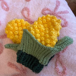 Könnte beinhalten: Handgemachte gehäkelte Maiskolben. Die Maiskörner sind gelb, die Blätter sind grün und die Basis ist dunkelgrün. Der Mais liegt auf einer rosa, strukturierten Oberfläche mit einem floralen Muster.