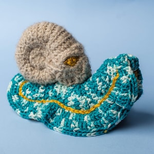 May include: A handmade crochet snail toy. The snail's shell is a light tan color, and the body is teal with white accents. A yellow line traces the snail's body. The snail's eye is yellow.