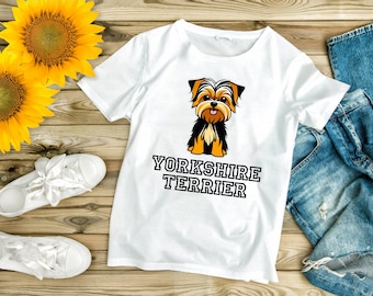 Op de afbeelding: Wit T-shirt met een cartoon Yorkshire Terrier illustratie in zwart, bruin en oranje, met de tekst "YORKSHIRE TERRIER". Het shirt wordt getoond met een blauwe jeans, witte sneakers en zonnebloemen op een houten oppervlak.