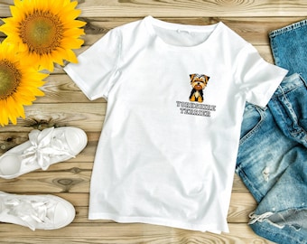 Op de afbeelding: Wit T-shirt met een cartoon Yorkshire Terrier en de tekst "Yorkshire Terrier" bedrukt op de voorkant. Het T-shirt ligt op een houten oppervlak met een paar witte sneakers, een zonnebloem en een blauwe jeans.