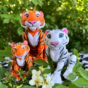May include: Three toy tiger figurines, one orange and white, one white with black stripes, and one orange with black stripes. The tigers are sitting on a green leafy surface.
