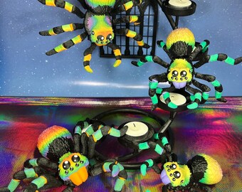May include: Four colorful, hand-painted, spider figurines with black legs and green and yellow accents. The spiders are displayed on a black metal candle holder.
