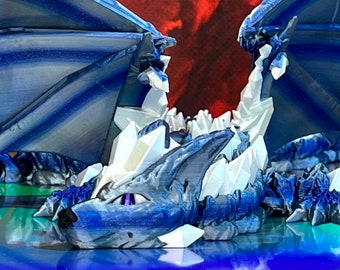 May include: A blue and white crystal dragon figurine with wings spread wide. The dragon is lying on its belly with its head raised and its eyes open. The figurine is made of a smooth, shiny material and has a detailed design.