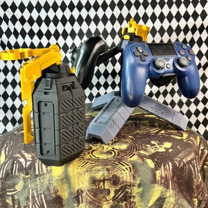 May include: A black and gold grenade-shaped phone holder and a blue and gold video game controller stand on a patterned surface.