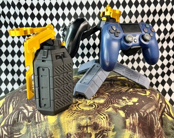 May include: A black and gold grenade-shaped phone holder and a blue and gold video game controller stand on a patterned surface.
