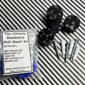 May include: A kit for mounting a snowboard to a wall. The kit includes two black wall mounts with a geometric pattern, a board latch, three black caps, three wall anchors, and three screws. The kit is packaged in a clear plastic bag with a white label that reads "The Ultimate Snowboard Wall Mount kit" and lists the kit contents. The label also includes the Etsy shop name and the phrase "Made in the USA."