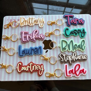 May include: A collection of personalized keychains with names like "Brittany," "Erika," and "Lauren." Each keychain features a gold-colored clasp and ring. The names are in various colors, including pink, blue, and red, with a layered design.