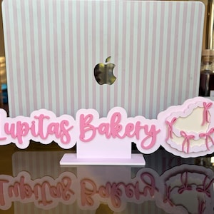 May include: A pink sign for "Lupitas Bakery" with a decorative design. The sign is made of layered pink material and features a cursive font. The background is a laptop with a pink and white striped pattern.