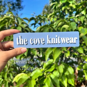 May include: A light blue rectangular sign with the text "the cove knitwear" in white raised letters.