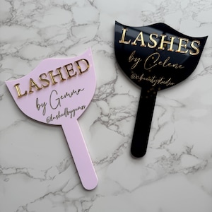 May include: Two pink and black acrylic signs with gold lettering. The pink sign says "Lashed by Gemma @lashedbygemma" and the black sign says "Lashes by Celene @celenebeautystudio".