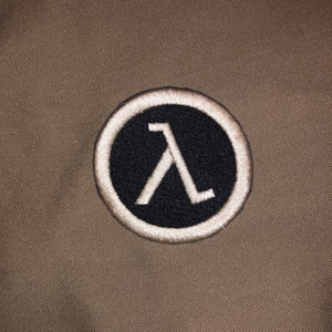 May include: A white embroidered patch with a black circle and a white Lambda symbol inside. The patch is on a brown fabric background.