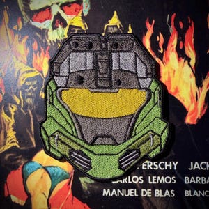 May include: An embroidered patch featuring a stylized green helmet with a prominent gold visor and dark grey detailing. The design includes a geometric structure with black borders, showcasing a detailed recreation of a futuristic combat helmet.