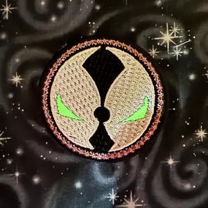 May include: Embroidered patch featuring the Spawn logo. The circular patch has a black center with a white and tan outer design, outlined in red. The eyes are bright green. The background is black with white stars and swirls.