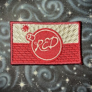 May include: A rectangular embroidered patch featuring a horizontal split design. The top half is off-white with a small red sun icon, and the bottom half is red with a brick pattern. A central red circle contains the white embroidered text RED.