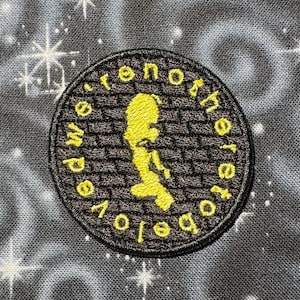 May include: A circular embroidered patch with the words "we're not here to be loved" in yellow around the edge. The center features a yellow silhouette of a person. The patch is on a black and white patterned background.