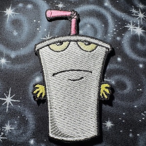 May include: Embroidered patch featuring a cartoon milkshake character with a grumpy expression. The character is gray with yellow eyes, yellow hands, and a pink straw. The background is a dark blue fabric with a galaxy design.