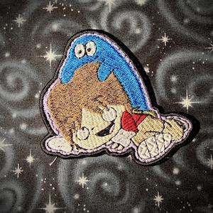May include: Embroidered patch featuring a cartoon character with brown hair, a red shirt, and a blue creature on top. The patch has a light purple border and is set against a dark background with white stars.
