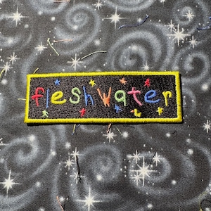 May include: A black iron-on patch with yellow stitching around the edges. The patch says "fleshwater" in colorful, embroidered letters.