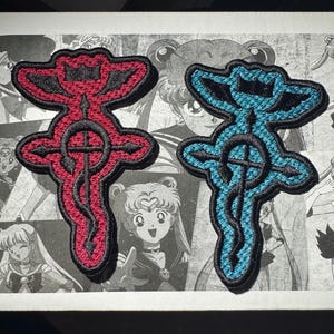 May include: Two embroidered patches, one red and one blue, with a black outline. Both patches feature a stylized design with a cross in the center.