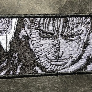 May include: A black and white embroidered patch featuring a close-up of a character's face with a furrowed brow and a determined expression.