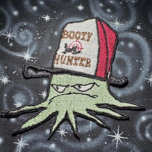 May include: Embroidered patch featuring a green octopus with an angry expression, wearing a hat that reads "Booty Hunter." The hat is white with red and black accents. The background is a dark, starry design.