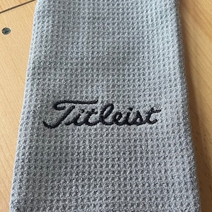 May include: Grey golf towel with a black embroidered "Titleist" logo. The towel has a waffle texture and a loop for hanging.