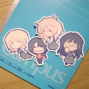 May include: Four cartoon stickers featuring anime-style characters with various expressions. The characters have different hairstyles and wear dresses or tops and pants. The stickers are on a blue notebook with Japanese text.