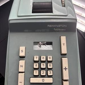 Könnte beinhalten: Ein vintage Remington elektronischer Taschenrechner in einem salbeigrünen Kunststoffgehäuse. Das Gerät verfügt über ein zentrales Tastenfeld mit weißen Tasten, einen Papierschlitz an der Oberseite und Funktionstasten mit S, N, -, T, +, X Symbolen.