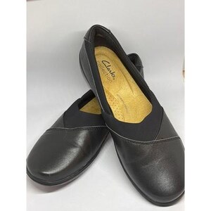 May include: A pair of black slip-on shoes with a rounded toe and elastic side panels. The interior footbed is tan and features the text Clarks Collection printed in black. The shoes have a smooth exterior finish and a low-profile design.