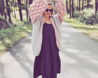 May include: A woman wearing a grey and pink knitted cardigan with large, puffy sleeves. She is wearing a black dress and black shoes. The cardigan has a loose fit and is open at the front. The woman is standing on a path in a forest.