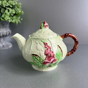 May include: A ceramic teapot with a light green leaf design and a brown handle. The teapot features a floral motif with pink and green accents. The lid is topped with a small floral detail. The teapot is a decorative item.