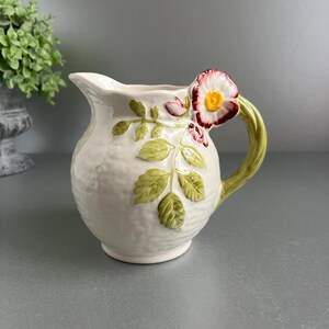 May include: Cream ceramic jug with a textured finish. The handle is green, and the jug is decorated with a red and white flower and green leaves. A small spout is on the left side of the jug.
