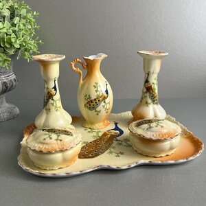 May include: A vintage porcelain vanity set featuring a peacock design. The set comprises a tray, two candle holders, two lidded jars, and a pitcher. The pieces are decorated with peacocks, flowers, and gold accents.