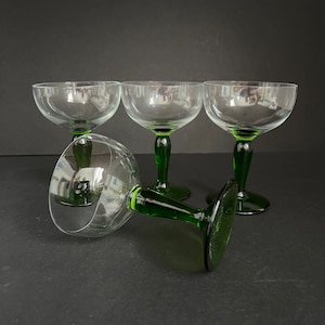 May include: Four clear glass champagne coupes with green stems. The glasses are arranged in a semi-circle on a black surface.