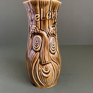 May include: A brown glazed ceramic vase featuring a relief design of a face with swirling eyes and a beard. The word Celery is embossed near the top rim. The surface has a textured, wood-grain appearance with darker brown accents in the grooves.