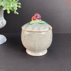 May include: A small, off-white ceramic jar with a lid, featuring a textured leaf pattern. The lid is decorated with a cluster of colourful berries and green leaves. The jar sits on a pedestal base, and is likely used for storing small items.
