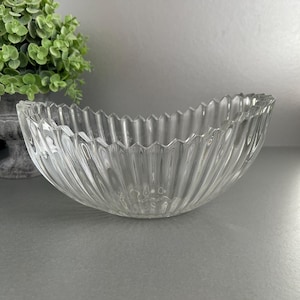 May include: A clear glass bowl with a fluted, scalloped rim. The bowl has vertical ridges, creating a textured look. The bowl is empty and rests on a grey surface. A green plant is visible in the background.