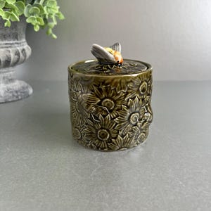 May include: An olive green ceramic jar with a lid, decorated with a sunflower pattern. The lid is topped with a bee-shaped handle with orange and yellow detailing. The jar is designed for storing items like sugar or small trinkets.