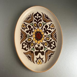 May include: Oval ceramic serving platter with a cream base and a brown, yellow, and green floral design. The platter has a brown rim and a symmetrical pattern in the centre. A decorative piece for serving food.