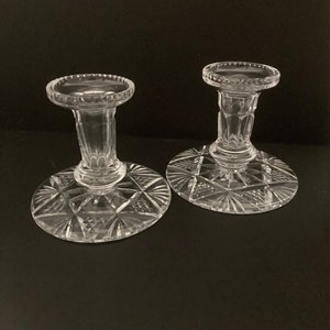 May include: A pair of clear glass candle holders with a cut glass design. The candle holders have a round base and a tall, slender column.