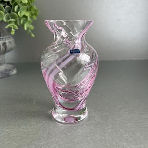 May include: A clear glass vase with pink swirls. The vase has a flared top and a rounded base. A small label is visible on the front. The vase is designed for decorative purposes.