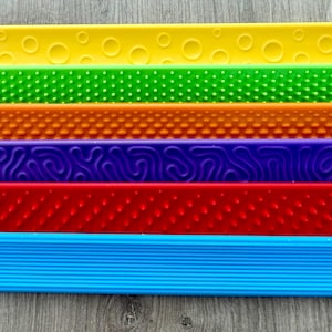 Könnte beinhalten: Eine Sammlung farbenfroher, strukturierter Sensorikstreifen in Gelb, Grün, Orange, Lila, Rot und Blau. Jeder Streifen weist ein einzigartiges taktiles Muster auf, darunter Punkte, Noppen und Linien. Die Streifen sind in einer parallelen Reihe angeordnet.