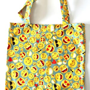 Könnte beinhalten: Eine blaue Tragetasche mit einem Muster aus gelben Smiley-Gesichtern mit verschiedenen Ausdrücken, darunter Sonnenbrillen, Herzen und herausgestreckten Zungen.